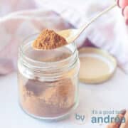 A square photo with a glass jar filled with shawarma spices. A pink white tea towel in the background. A spoon scoops the mix out of the jar.
