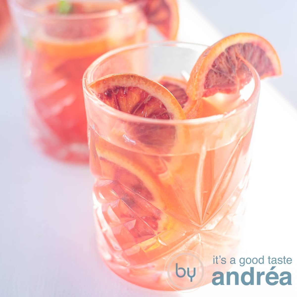A square photo with two glasses filled with blood orange iced tea, orange slices and ice on a white background