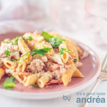A square photo with a pink plate of pasta with cherry tomatoes and tuna and parsley. A pink background.