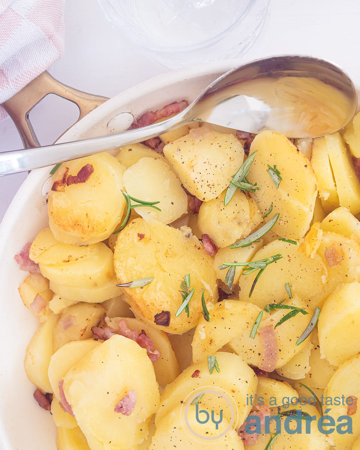 A photo from above of a white frying pan with fried potatoes, onion, rumarine and bacon bits. A spoon scoops it in.