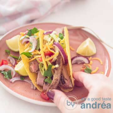 A square photo with a pink plate with tacos filled with sirloin fresh vegetables and topped with Cheddar cheese. A hand holds a taco
