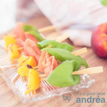 A square photo with a glass dish with tapas sticks with grilled peach, ham and basil on a wooden background.