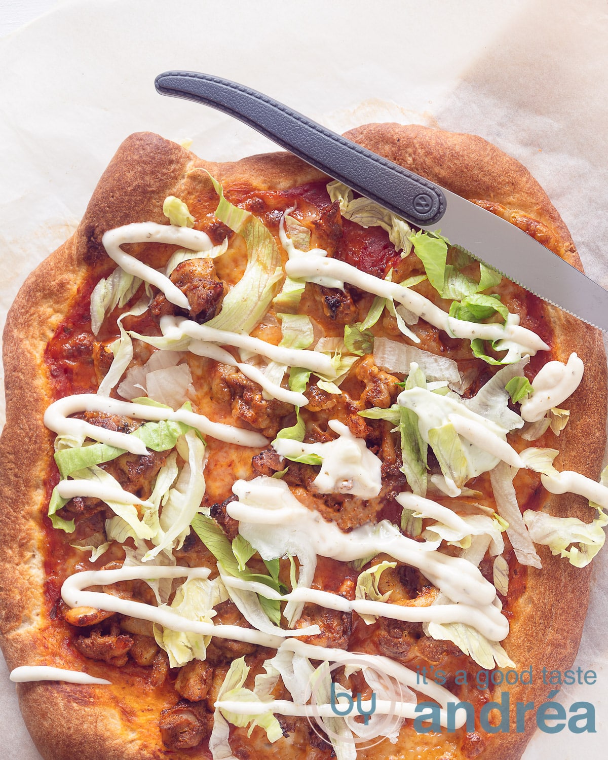 A photo from above of a pizza with chicken kebab, iceberg lettuce and a drizzle of garlic sauce. A knife rests on top. A white background.