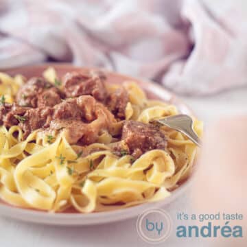 A square photo with a pink plate with pasta and beef in a creamy sauce. A hand pierces the flesh. A pink white tea towel in the background.