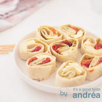 A square photo with a round white plate filled with 8 caprese wrap snacks. A white background. In the background a pink and white tea towel.