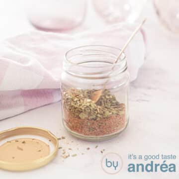 A square photo with a glass jar filled with gyros spice mix in the middle. A golden spoon is in it. The lid is next to it. A pink white tea towel and pink glasses in the background.