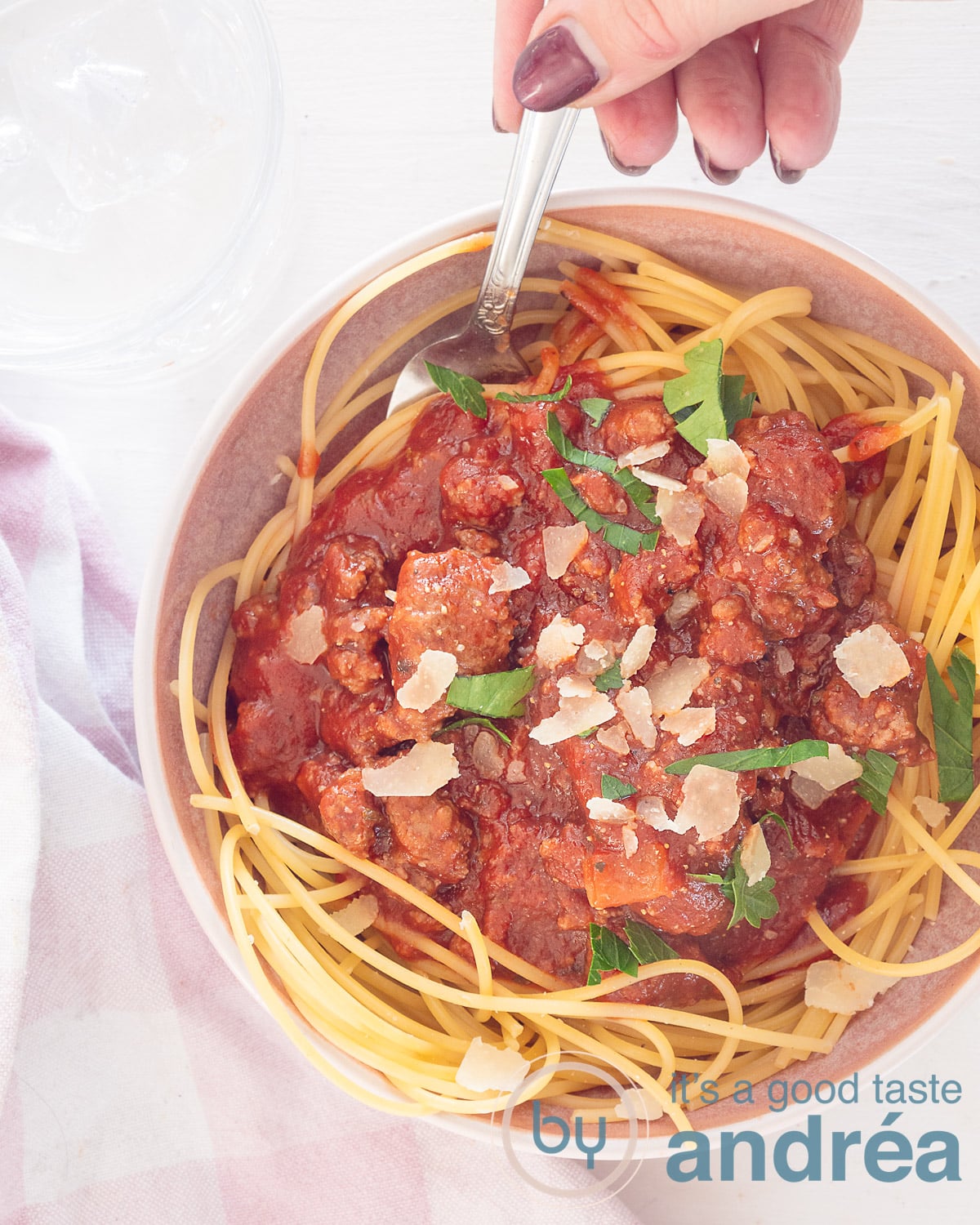 A photo from above of a bowl of spaghetti topped with homemade bolognaise sauce with Parmesan cheese and parsley. A hand holds a spoon. A white background, at the bottom left a white pink tea towel.