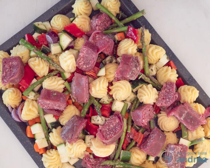A baking tray, not yet baked, with pommes duchesse, various vegetables and pieces of beef finch on a white surface.