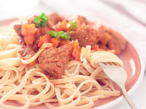 A square photo with a pink plate with tagliatelle and Italian stew
