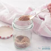 A square photo with a glass jar with chicken spices that are mixed themselves. A spoonful of mix is scooped out. A pink white tea towel in the background.