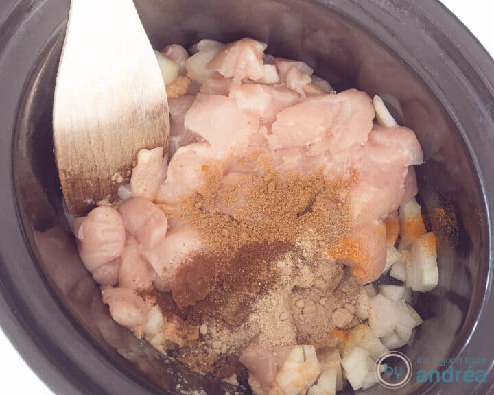 A slow cooker filled with chicken and herbs. A wooden spoon sticks in it.