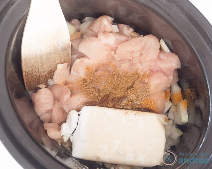 A slow cooker with chicken, herbs and coconut. A wooden spoon sticks in it.