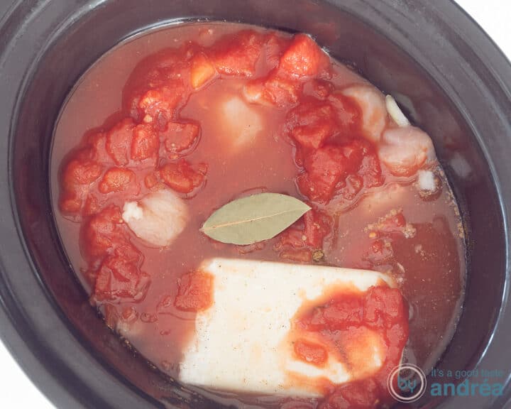 A slow cooker with chicken, herbs, coconut and diced tomatoes. A bay leaf floats in the center.