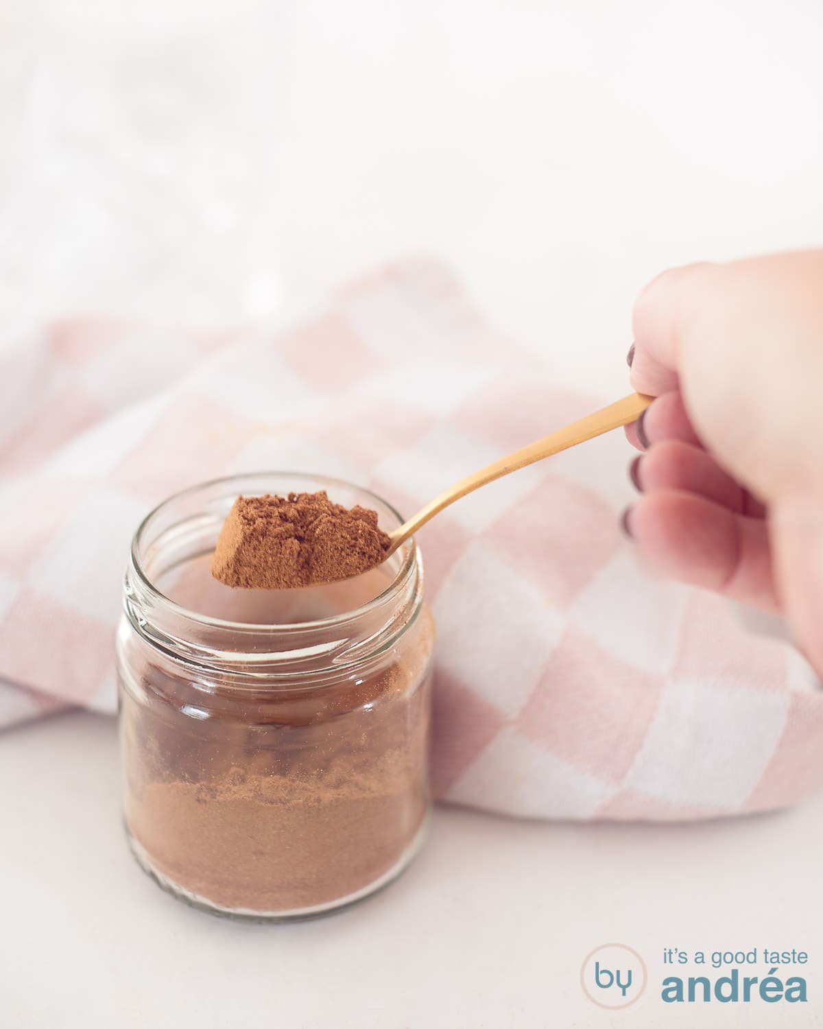 A glass jar with gingerbread spices with a golden spoon that has some mix on the spoon. A white background. In the background a pink and white tea towel