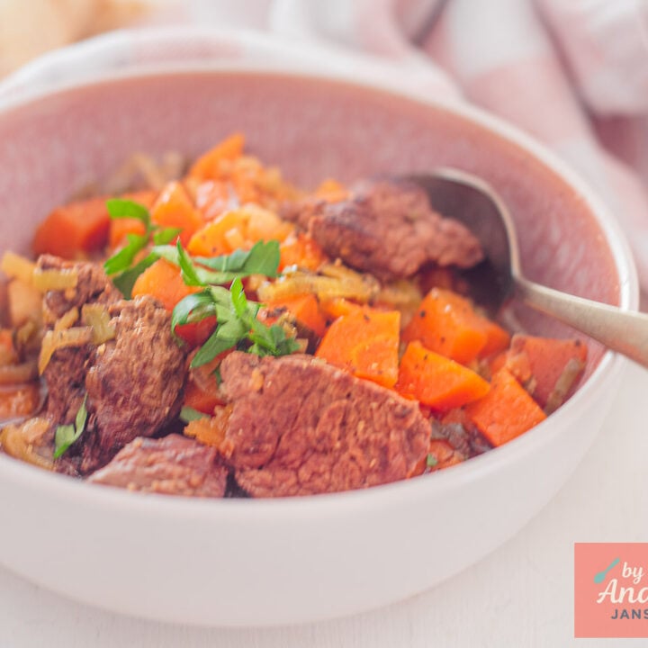 Roze kom met runderstoof uit de slowcooker, met stukken rundvlees en wortel in biersaus, peterselie en een lepel rechts.