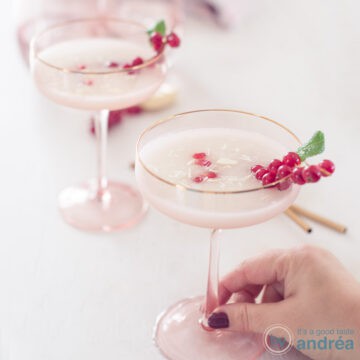 A square photo with two pink champagne glasses filled with scroppino, pomegranate seeds, red berries and mint. A hand grabs the front glass.