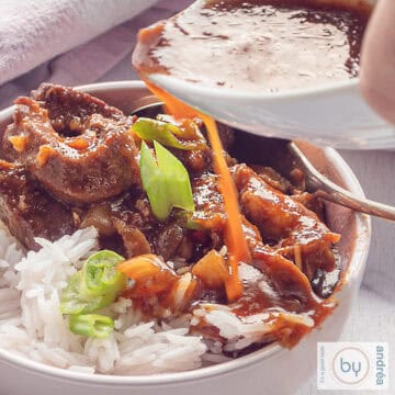 A square photo with a bowl filled with babi pangang, rice and a garnish of spring onions. A white om with sauce pour extra sauce over the meat