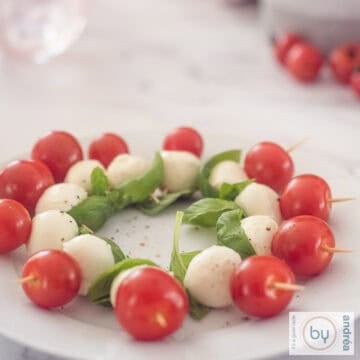 A square photo with a bowl filled with mini Caprese sticks with cherry tomatoes, mozzarella and basil leaves.