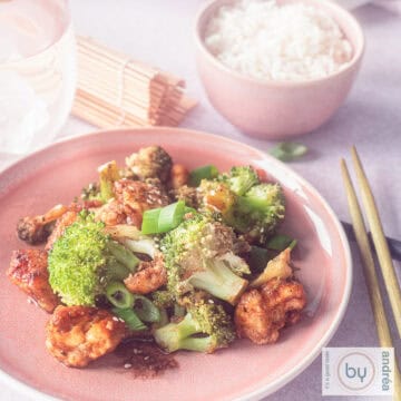 A square photo of a pink plate filled with broccoli, chicken and hoisin sauce. A bowl of rice in the background.