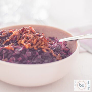 A square photo of a pink bowl filled with red cabbage and a topping of bacon on a white background.