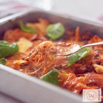 A square photo of a roasting pan filled with cheesy one-pan lasagna with mascarpone and minced meat. A spoon takes a bite.
