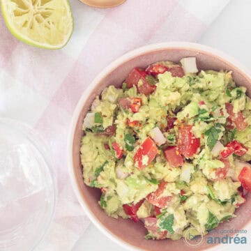 A square photo taken from above. Bottom right a bowl with homemade guacamole with avocado, tomato and lime. At the top a piece of lime is still visible.