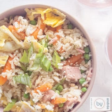 A square photo of a pink bowl with fried rice, vegetables, egg and ham on a white background.