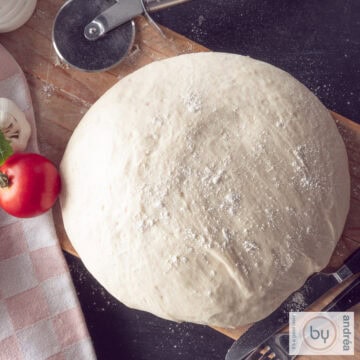 A square photo taken from above of a ball of pizza dough on a wooden board. A tomato and garlic and a pizza cutter are next to it.