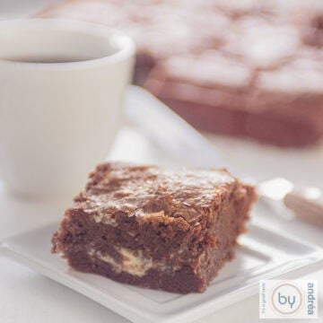 A square photo of a white plate with a piece of plate with melted white chocolate inside. A cup with coffee behind it together with brownies.