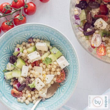 A square photo with two bowls, a blue one and part of a glass bowl filled with salad of pearl couscous, cucumber, onion, feta and tomatoes. A bunch of tomatoes above.