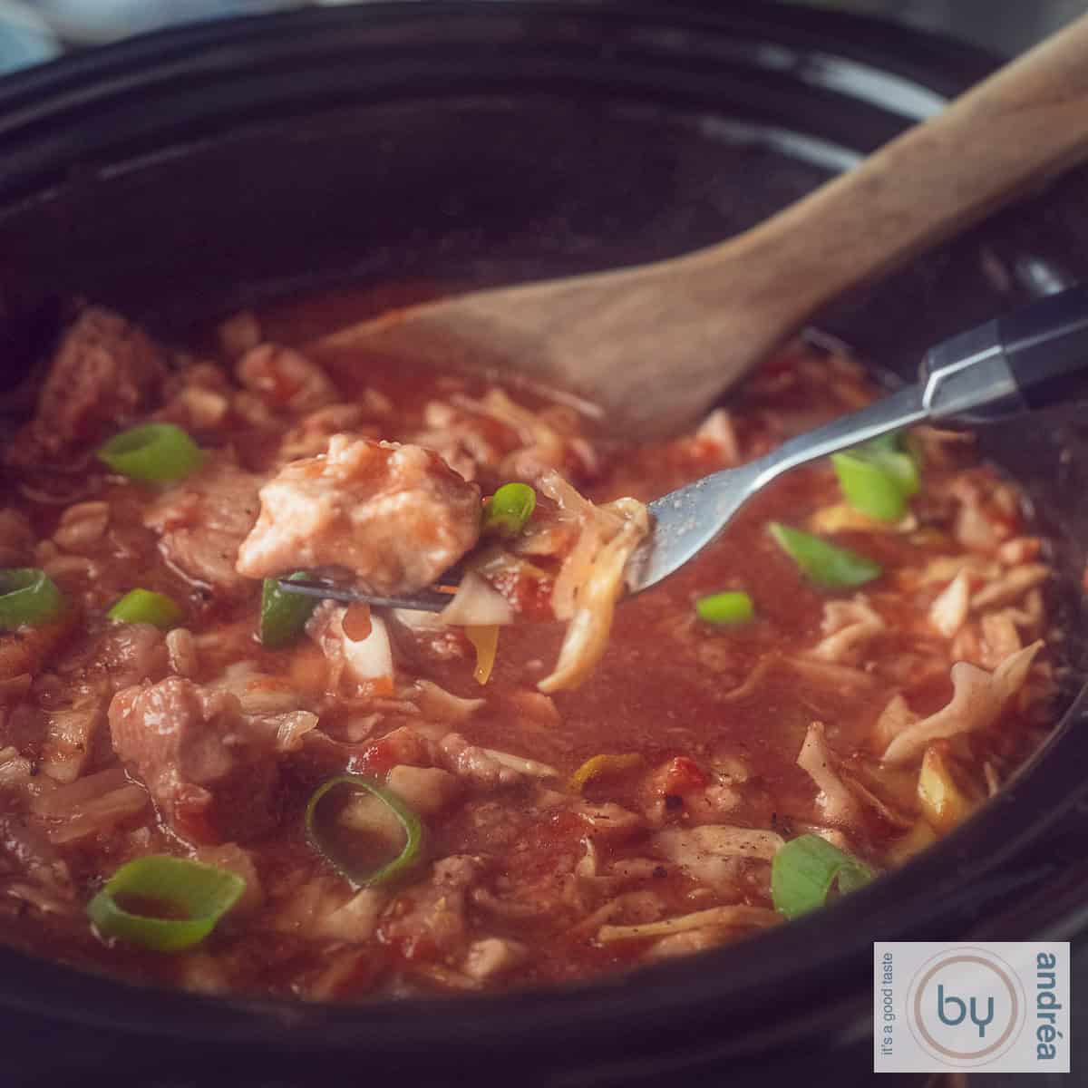 A square photo of part of a slow cooker filled with cabbage, ham steaks and a rich sauce. A fork picks a piece of meat from the pot.