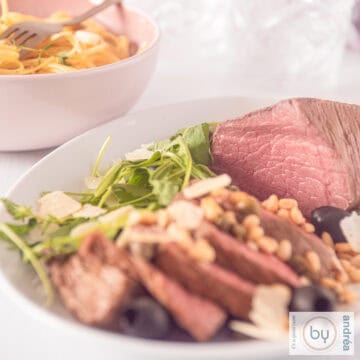 A square photo of roast beef with five slices cut off and garnished with pine nuts, olives, cheese and arugula. Linguine in the background.