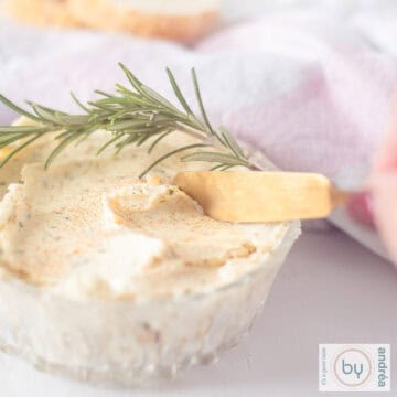 A square photo of a glass bowl with rosemary, butter, garlic and herbs. A knife scrapes off a little.