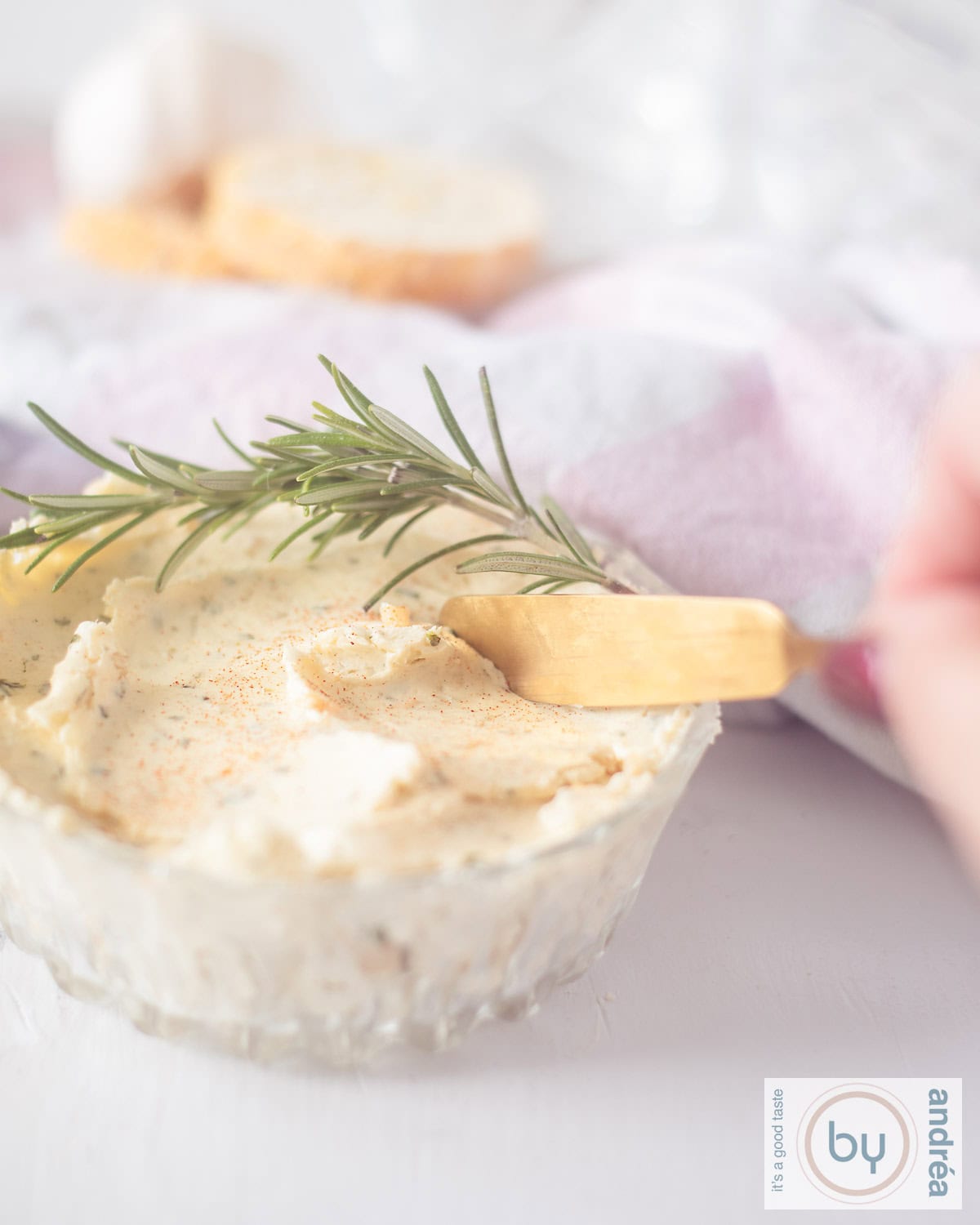 An aerial photo of a glass bowl with butter seasoned with herbs and Parmesan cheese. A gold knife scrapes some butter.