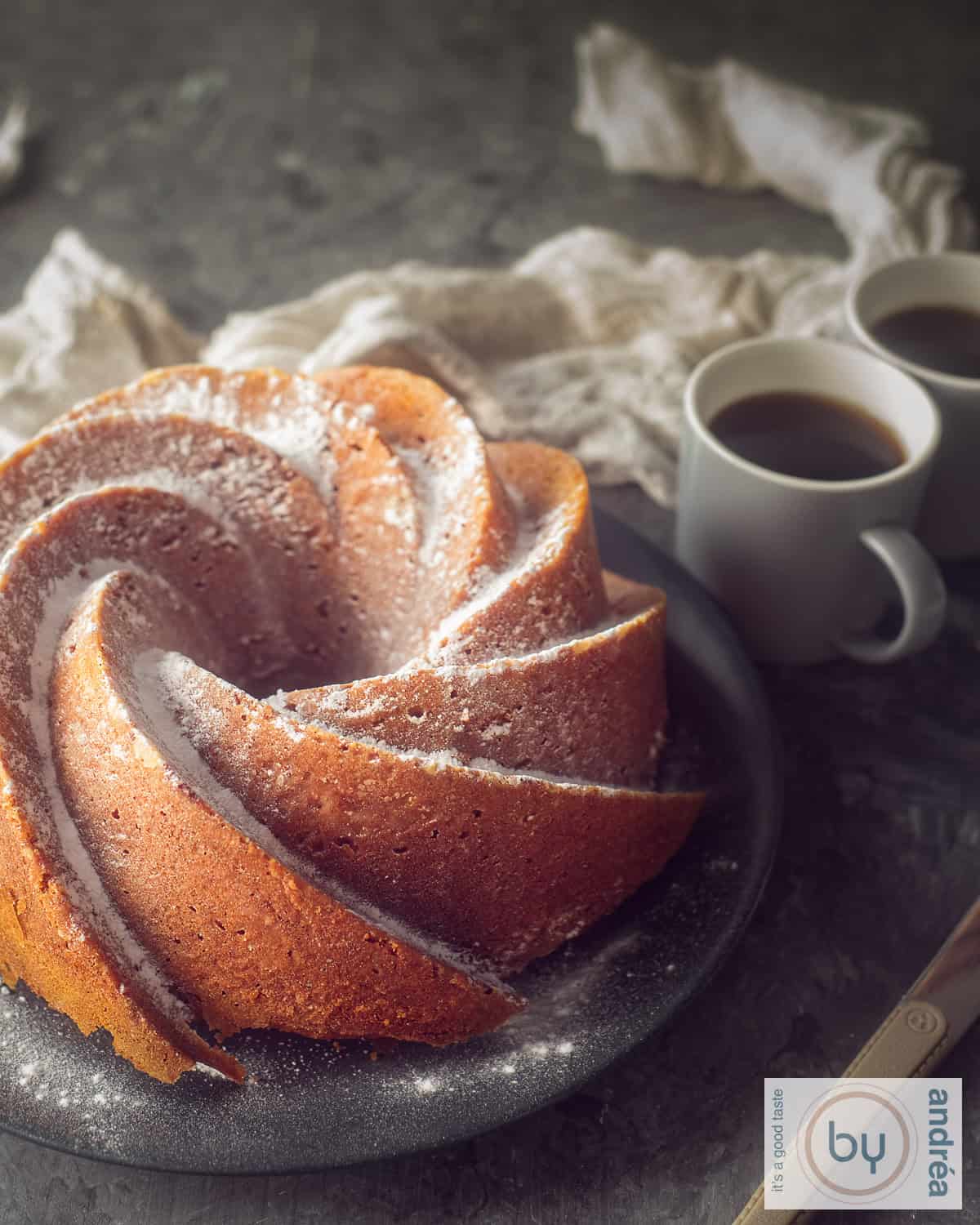 An elevation photo of a pumpkin bundt cake on a gray plate. Two cups of coffee behind it, a dark background.