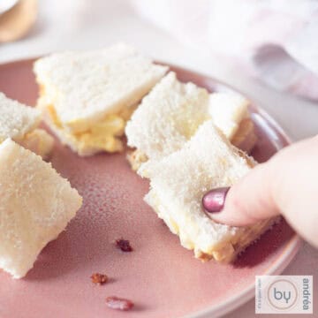 A square photo showing part of a pink plate with sandwiches with smoked chicken curry salad with bacon and egg. A hand takes a sandwich.