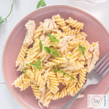 A square photo of a pink plate with pasta, creamy sauce with Parmesan and garlic and parsley on a white background