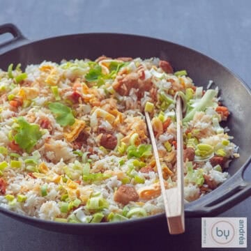 A square photo with a wok filled with nasi goreng with ham, chicken and bacon and vegetables. A wooden pair of tongs is in the wok. A dark background