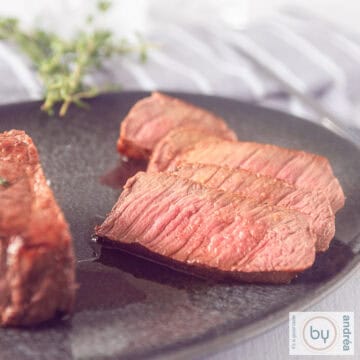 A square photo of roast beef, with slices cut off on a grey platter. Fresh herbs and a grey white cloth in the background.