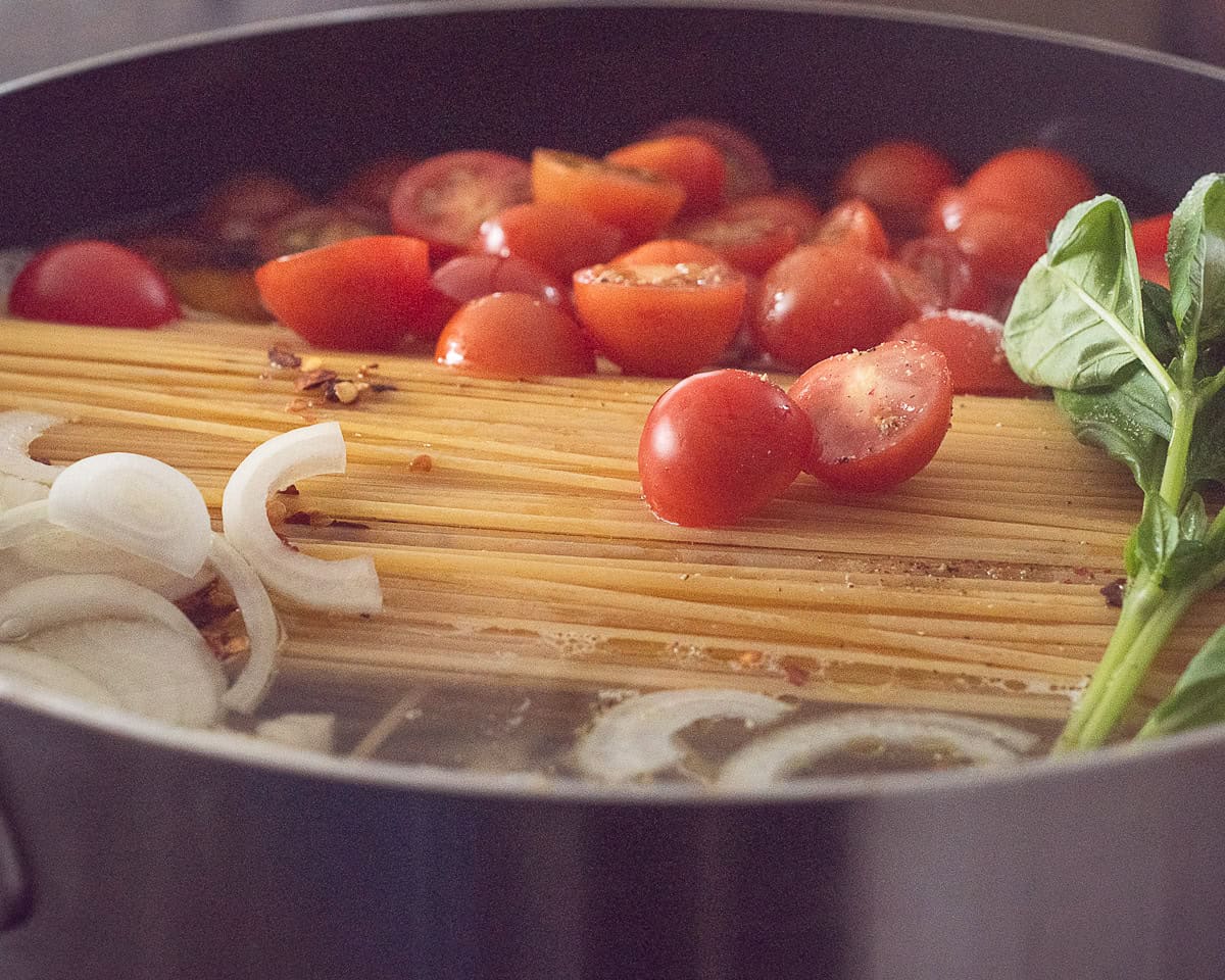 A casserole pan filled with pasta, onion, tomatoes, basil and stock.