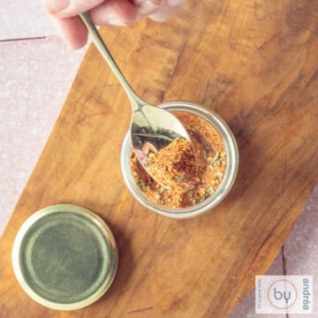 A square photo from above, a glass jar with Mexican spices. A golden spoon scoops out a bite.