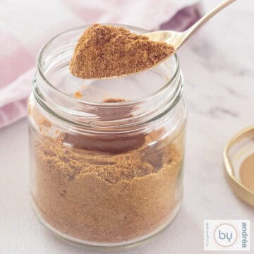 A square photo with a glass jar filled with curry madras spices. A spoon has grabbed a scoop.