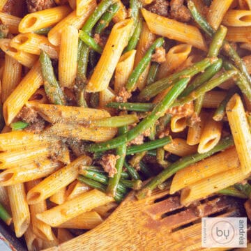A square photo of a pan filled with penne alla vodka, with green beans and minced meat. A wooden spoon is in the pan.