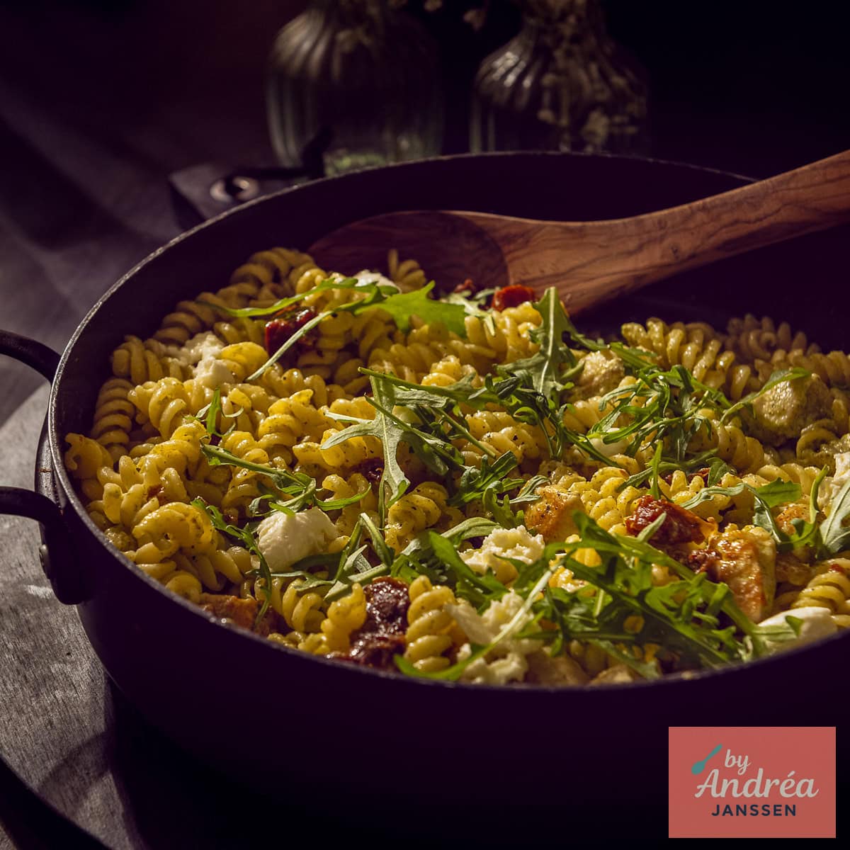 A square photo with a black pan of pasta pesto with chicken, arugula, and sun-dried tomatoes.