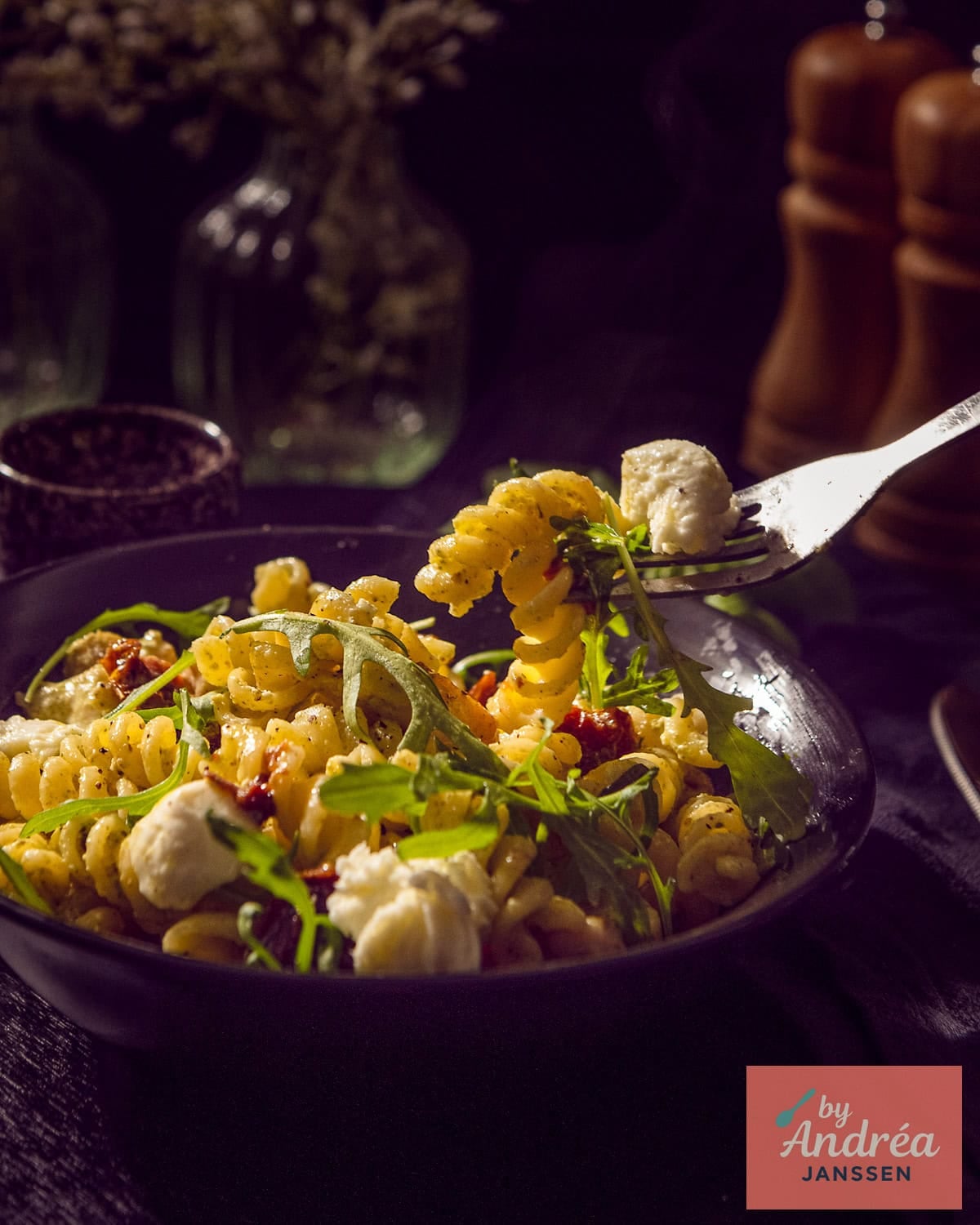 A small bowl of pasta with pesto sauce, arugula, mozzarella, sun-dried tomatoes, and chicken. A fork takes a bite.