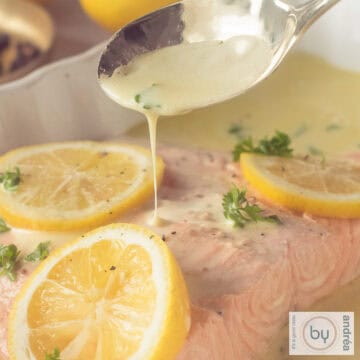 A square photo of a piece of salmon with lemon slices, parsley and white wine sauce. This is poured onto the fish with a spoon.