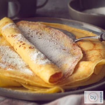 A square photo with a dark gray plate with a stack of freshly baked pancakes with powdered sugar.