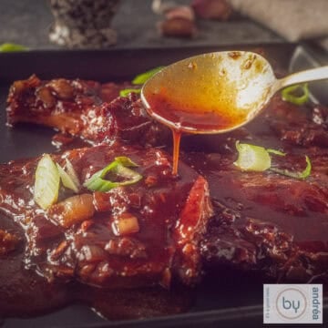 A square photo of pork chops with soy sauce. A spoon of sauce drips onto the chops.