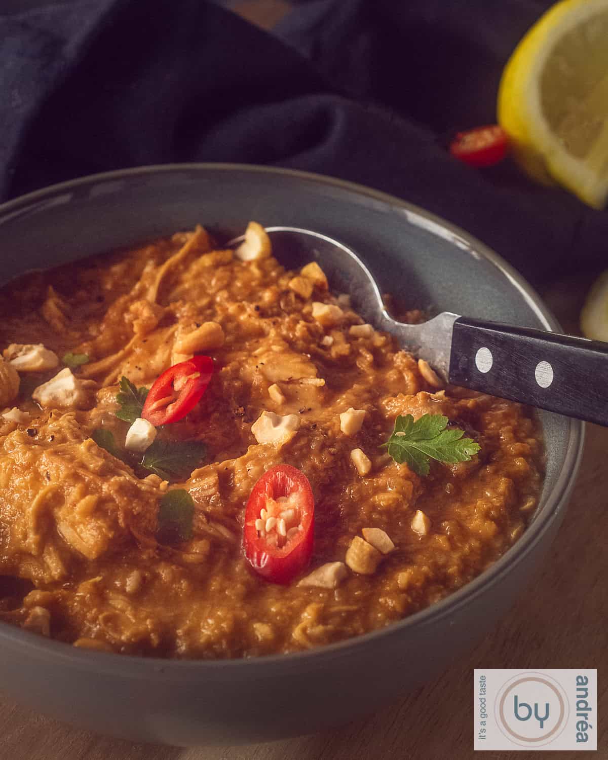 A grey bowl with a spoon containing peanut sauce with chicken pieces. Garnish of red pepper and parsley and nuts.