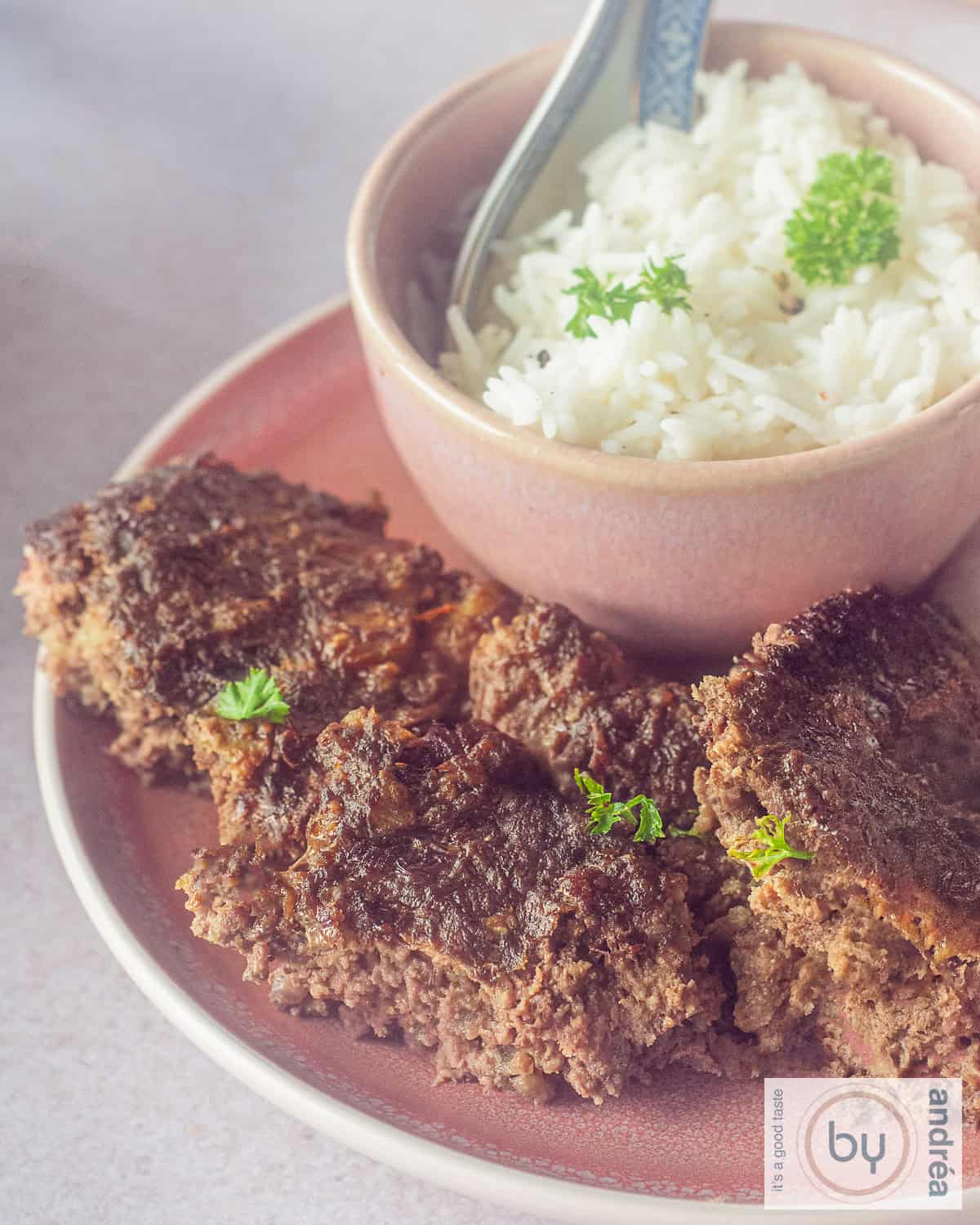Part of a plate with pieces of frikadel pan and a small bowl of rice.
